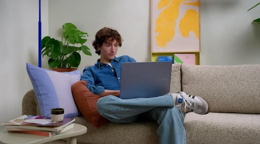 Jovem sentado em um sofá, usando um notebook com bateria de longa duração, vestido com camisa jeans.