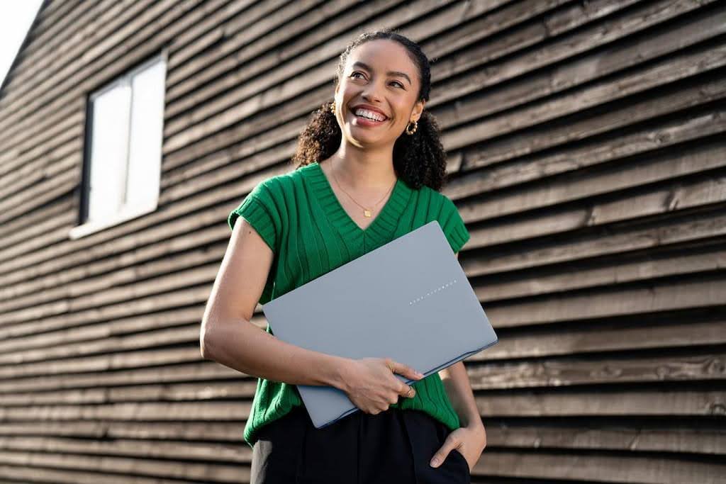 Uma mulher de cabelo cacheado e blusa verde, sorrindo enquanto segura seu notebook ASUS que ganhou de presente Dia das Mães.