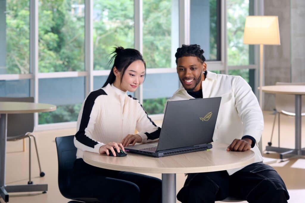 Dois jovens sorrindo enquanto trabalham juntos em um laptop ASUS em uma mesa, com janelas grandes ao fundo.