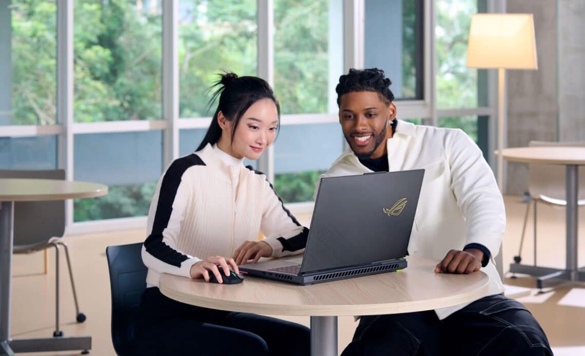 Dois jovens sorrindo enquanto trabalham juntos em um laptop ASUS em uma mesa, com janelas grandes ao fundo.