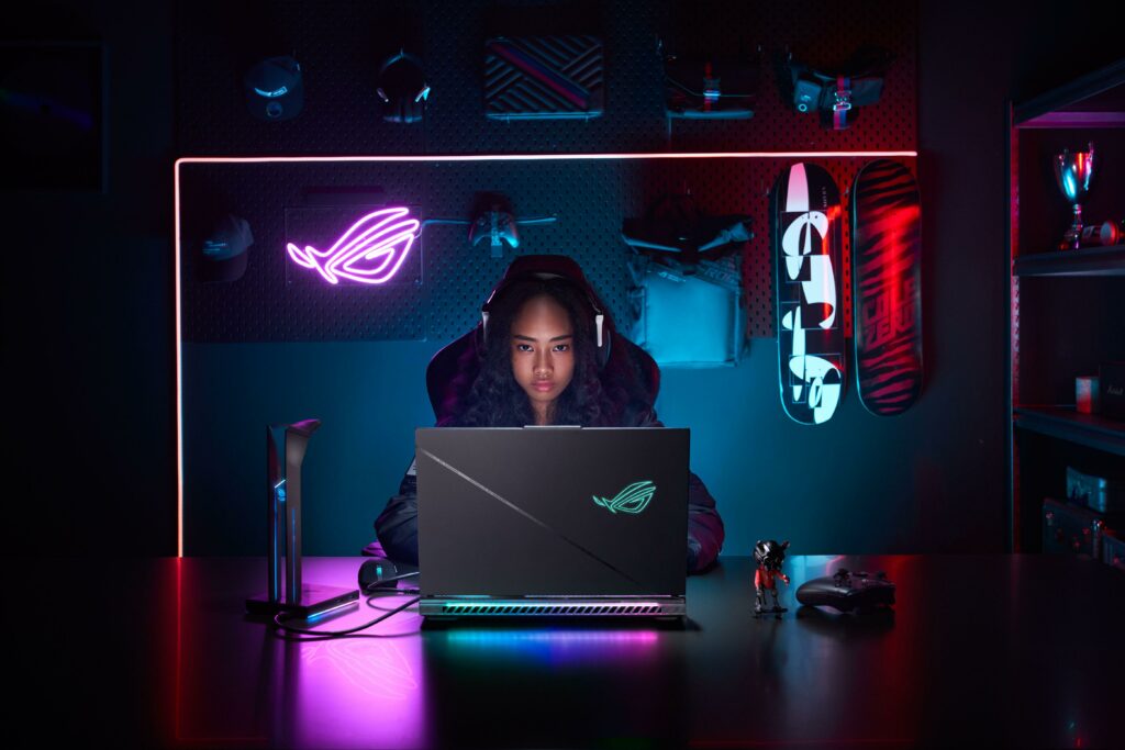 Jovem usando um notebook com iluminação neon em um ambiente moderno, com destaque para o ASUS ROG Strix Scar 18, ideal para jogos de alta performance.