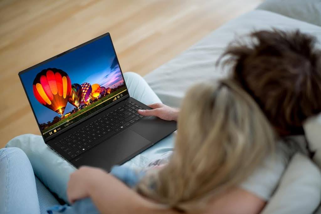 Duas pessoas abraçadas e sentadas no sofá, vistas por cima, assistem a um vídeo de balões coloridos ao pôr do sol em um notebook ASUS.