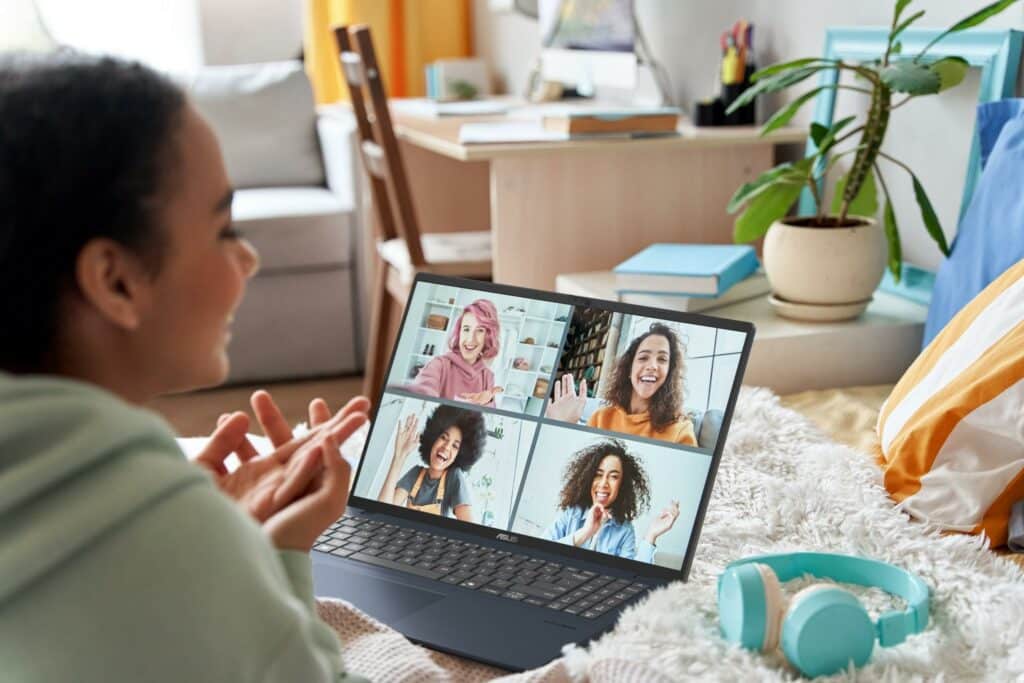 Mulher em casa usando o notebook ASUS Vivobook 16 para videoconferência, com amigas sorridentes na tela e ambiente confortável ao redor.